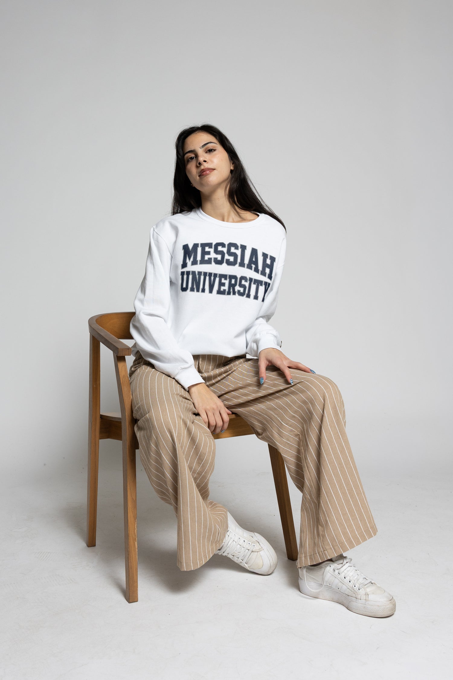 Woman wearing a 'Messiah University' sweatshirt sitting on a wooden chair against a plain background